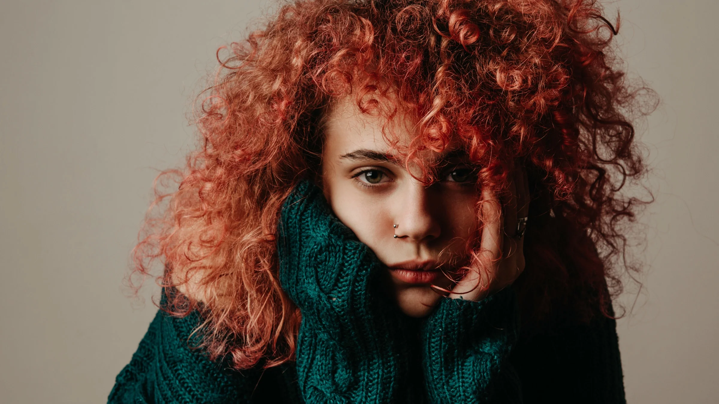 Portrait of a young woman with red curly hair with her face resting in her hands. She's wearing a green wool sweater and has a sad expression on her face.