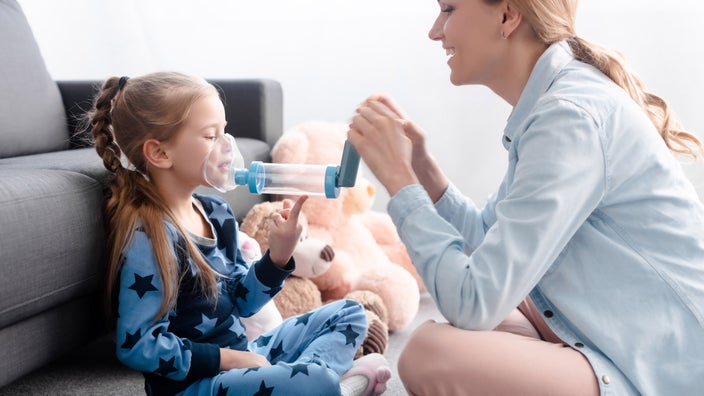 A parent and child sitting on the floor, using an inhaler with a spacer, surrounded by stuffed animals.