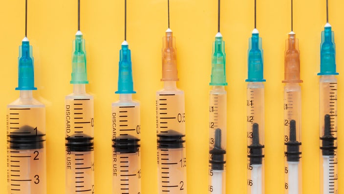 Eight syringes on a yellow background. Each syringe has a different color top tip. Some are blue, some are orange, and some are green.