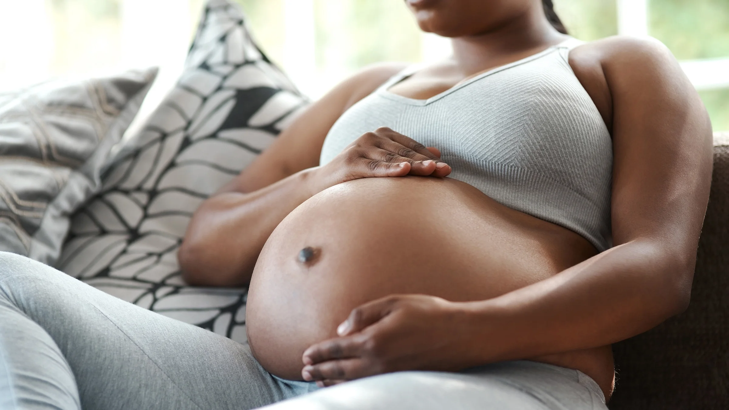 Cropped shot of a pregnant person holding their bare belly while they sit on the couch.