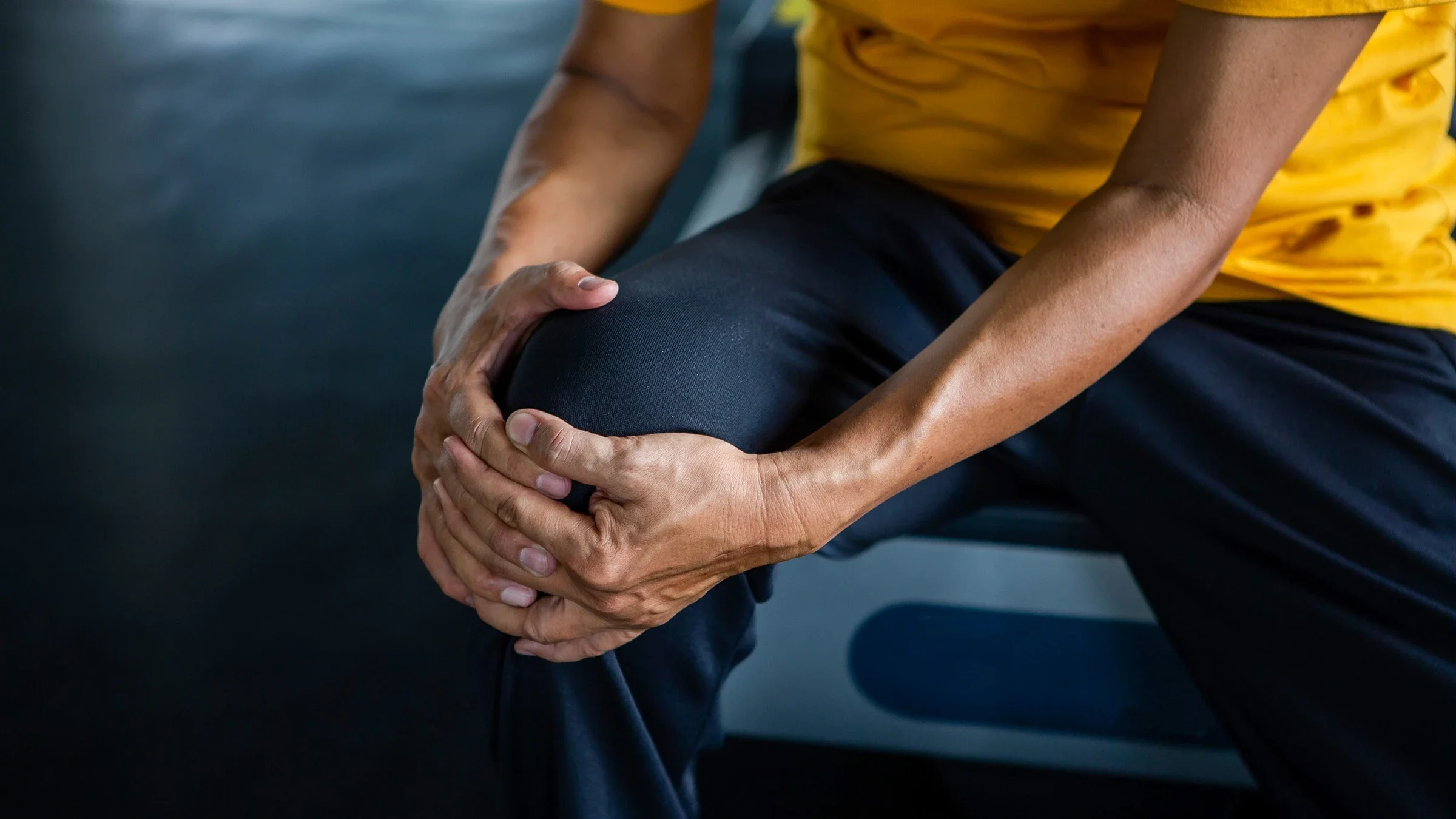 Close-up of a man holding his knee from arthritis pain. He is wearing sweatpants and a yellow t-shirt.