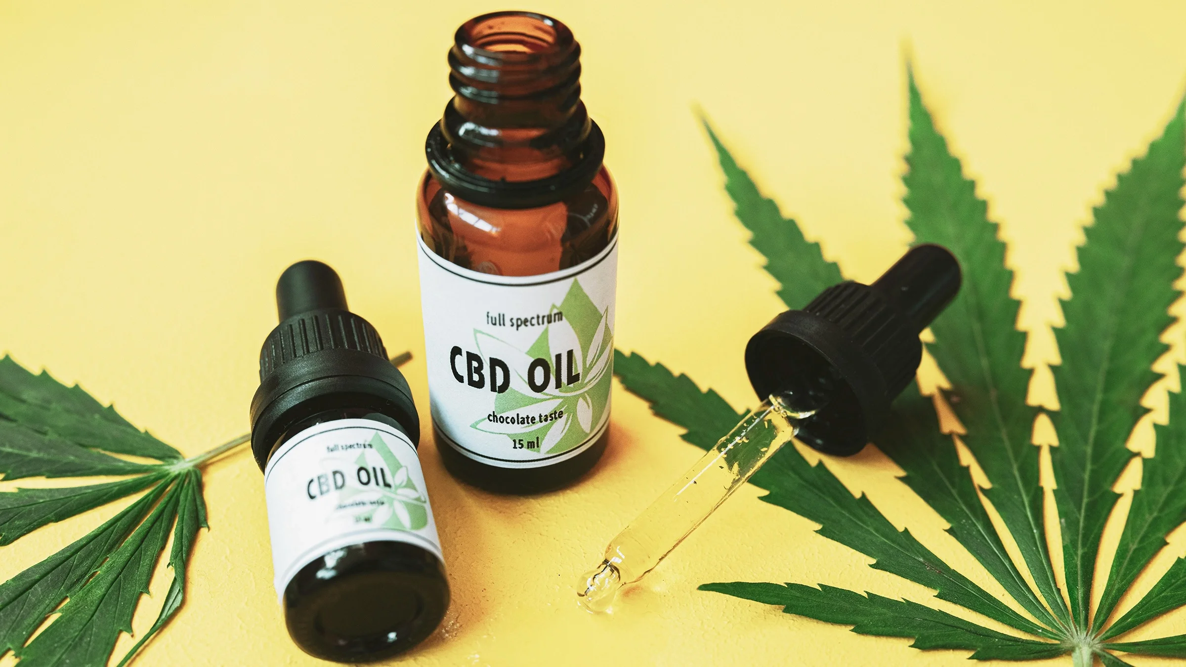 CBD oil bottles on a yellow background. One of the bottles has the dropper next to its side and there are two marijuana leaves in frame.