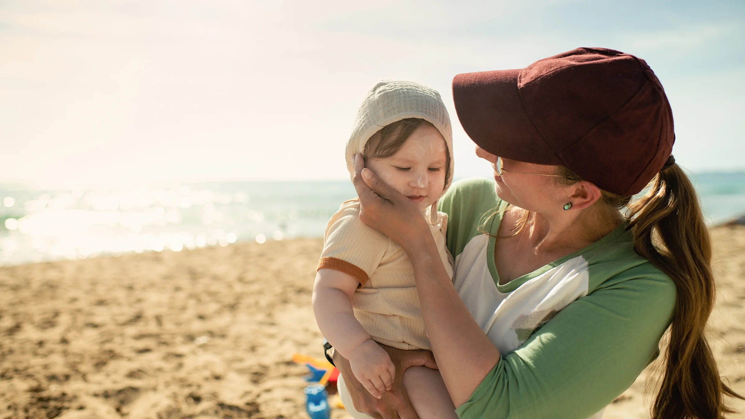 A woman applies sunscreen to her baby’s cheek while walking on the beach.