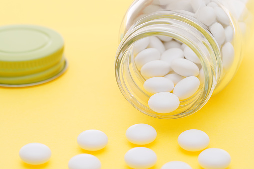 Glass bottle of white round pills spilling out on a yellow background.