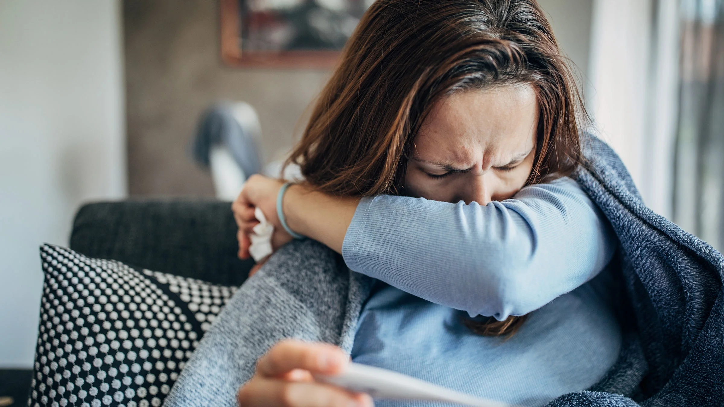 Woman with a cough sitting on sofa and holding thermometer