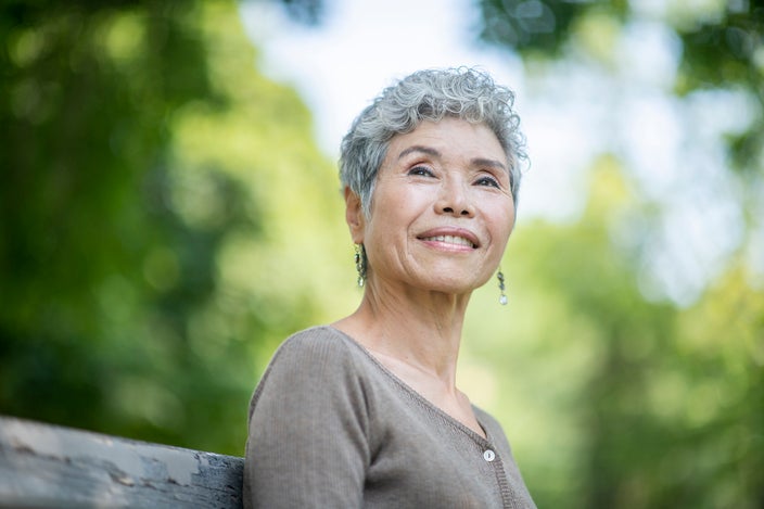 Portrait of an elderly woman with short gray hair. She is smiling softly as she sits on a bench outside in a park.