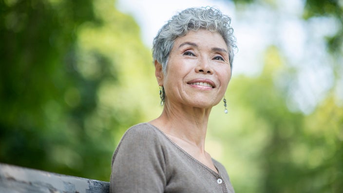 Portrait of an elderly woman with short gray hair. She is smiling softly as she sits on a bench outside in a park.
