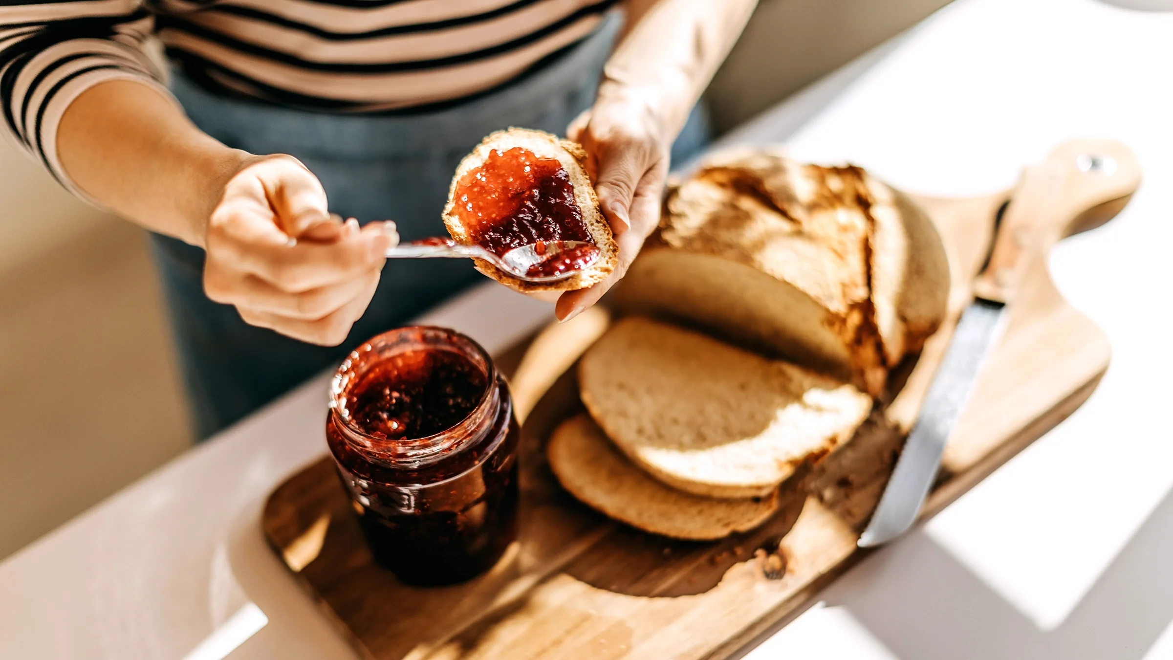 Cropped image of a person putting jelly on a piece of toast. There is a loaf of bread sliced on the cutting board next to them.