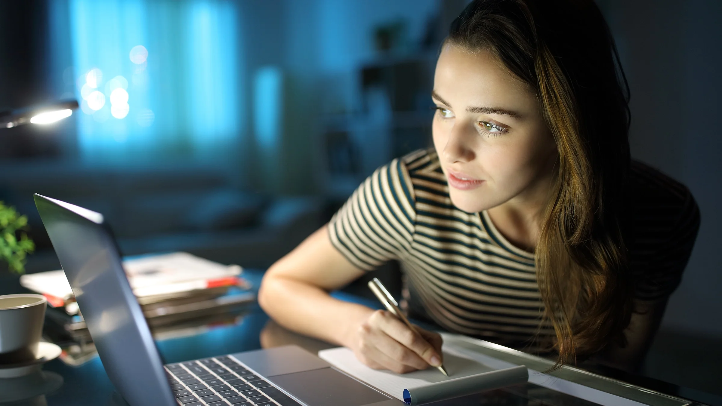 A woman writes notes while researching on a laptop.