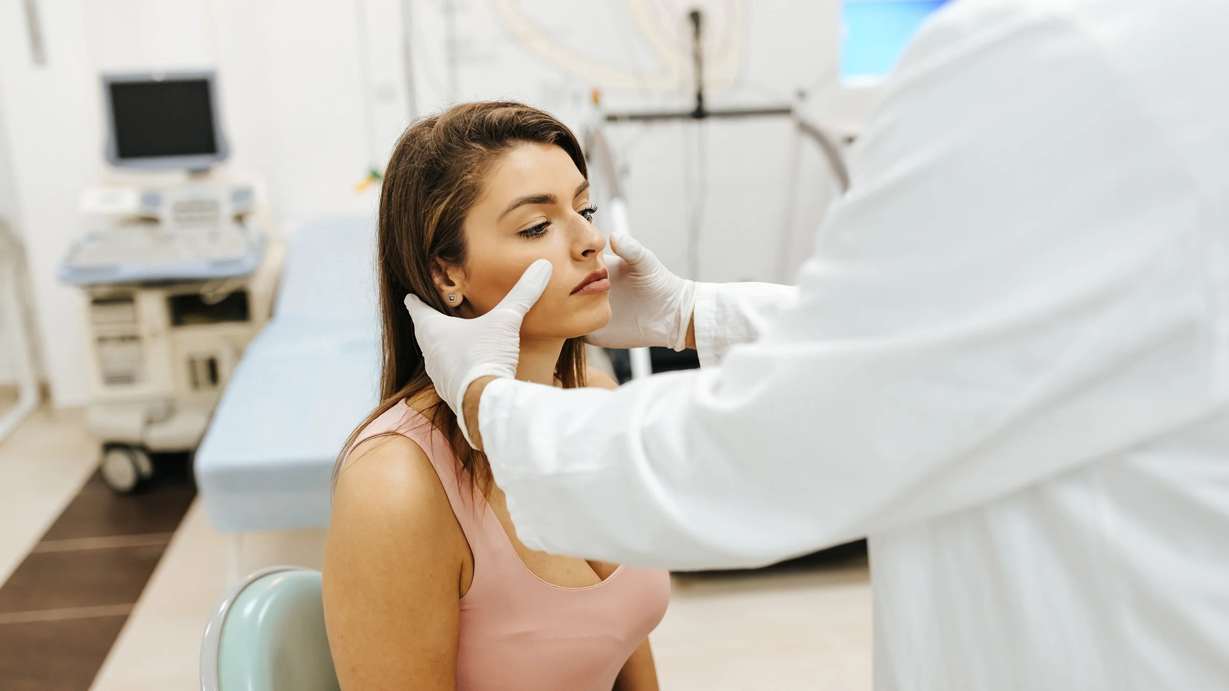 Young woman having her sinuses examined by her doctor. She is sitting in a chair next to a hospital bed.