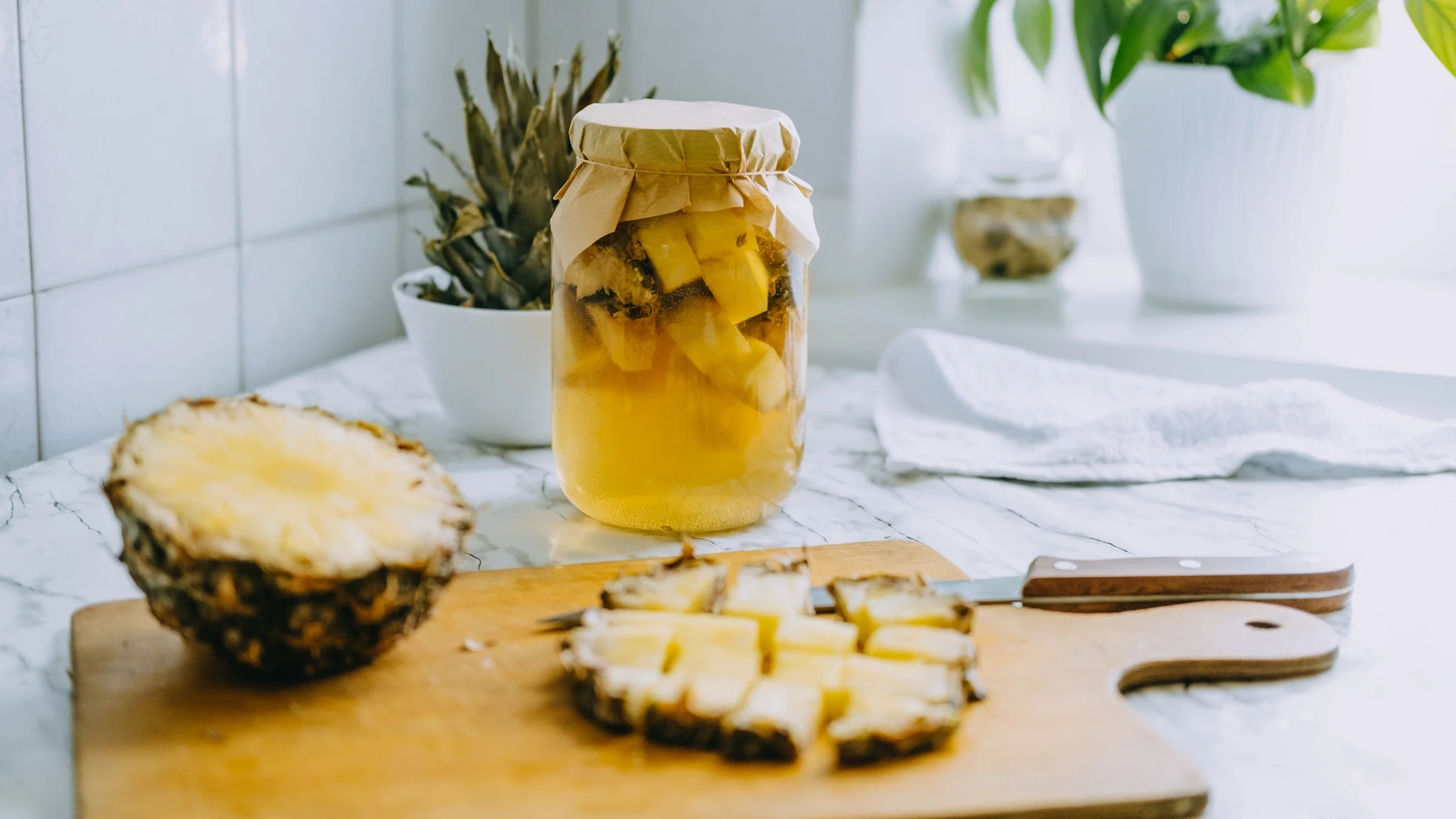 Jar of pineapple kombucha fermenting on a white marble tabletop. There is a cut open pineapple in front of the jar on a cutting board.