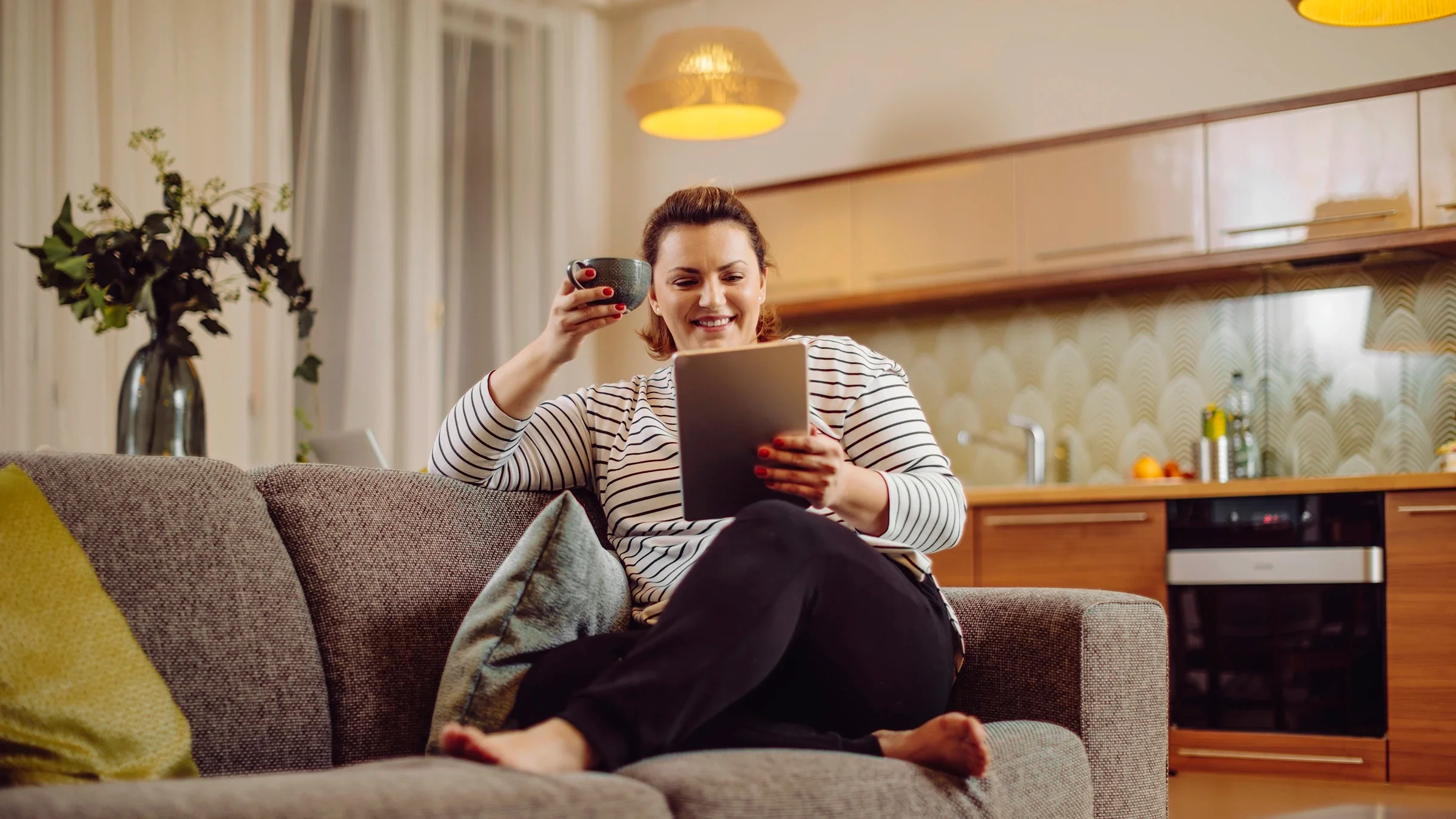 A woman drinks tea and reads her tablet.