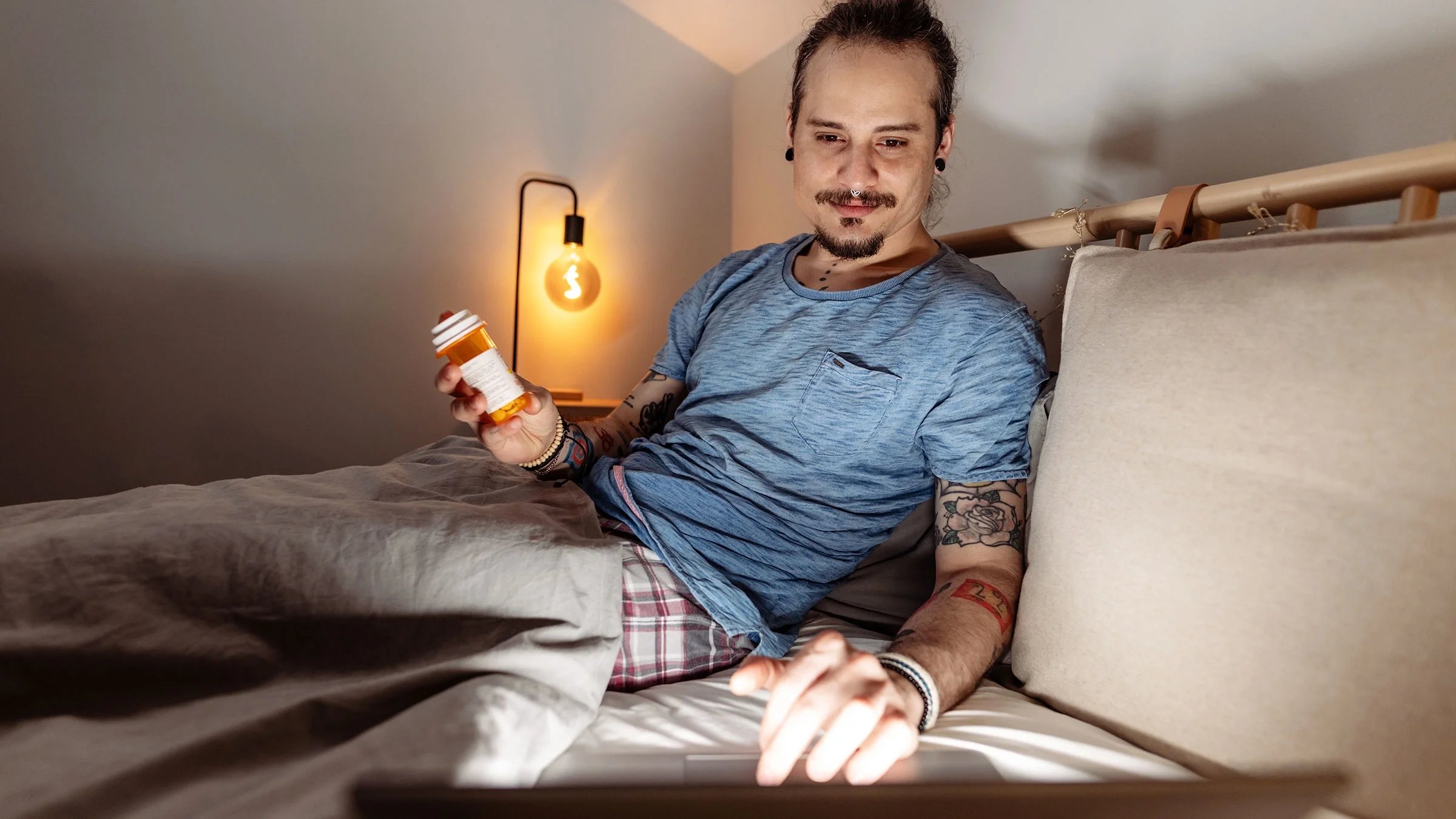 A man is sitting in bed and holding a pill bottle, while looking up the medication online.