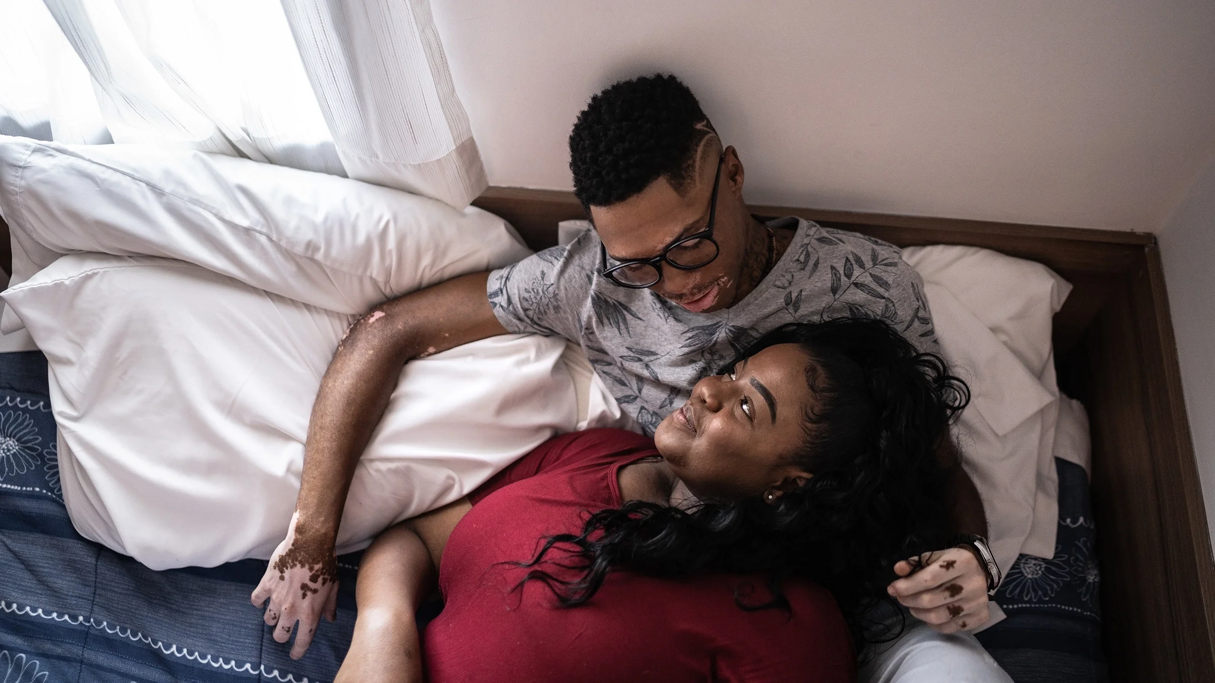Overhead shot of a couple cuddling up in bed. The woman is laying her head in the man's lap looking up at him lovingly.