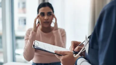 Young female patient talking to her doctor while showing where her pain is on her temples. The doctor's hands and clipboard are in focus while the patient is out of focus.
PeopleImages/E+ via Getty Images