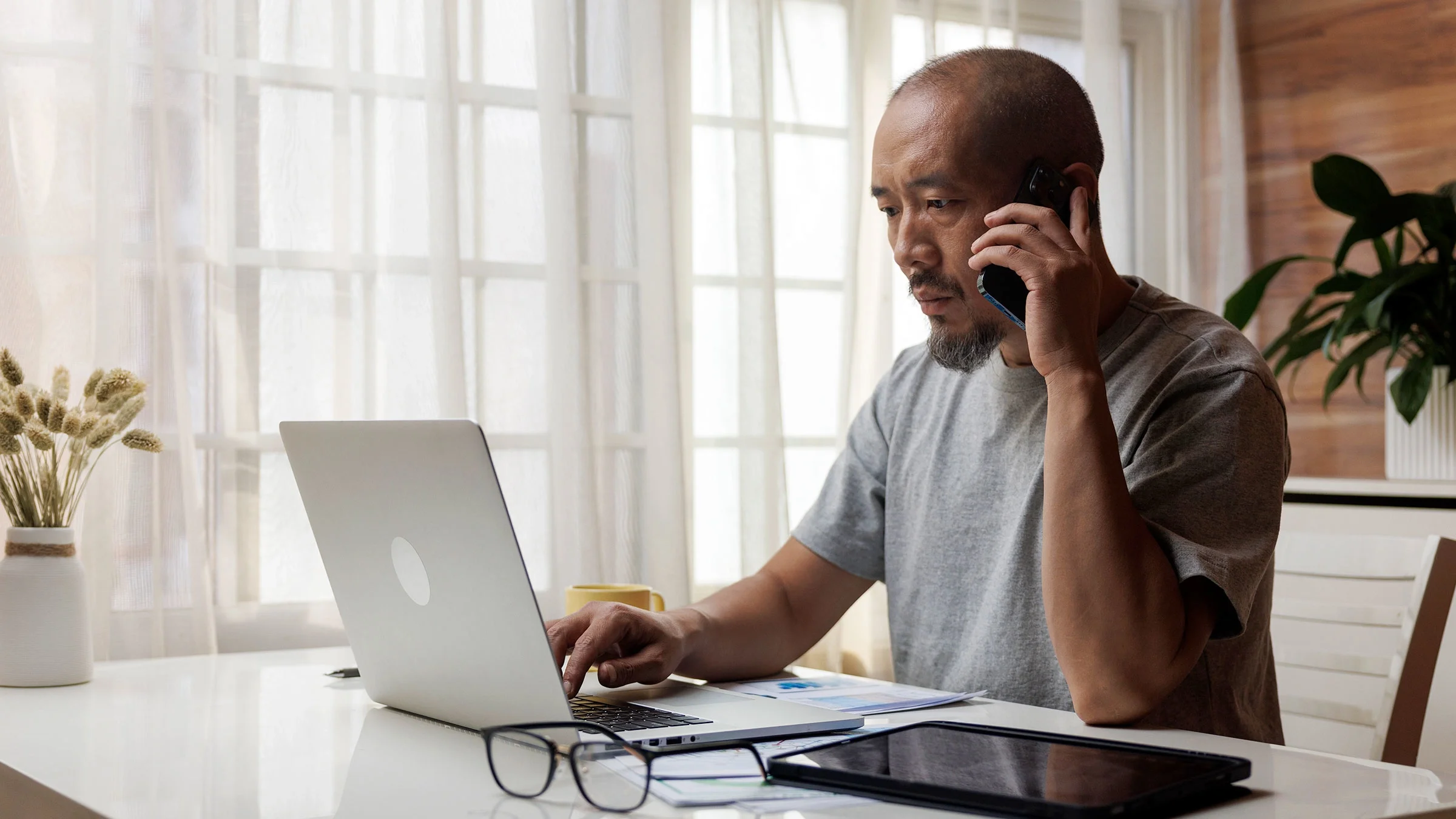 A man talks on the phone while sitting at a table working on his laptop.
