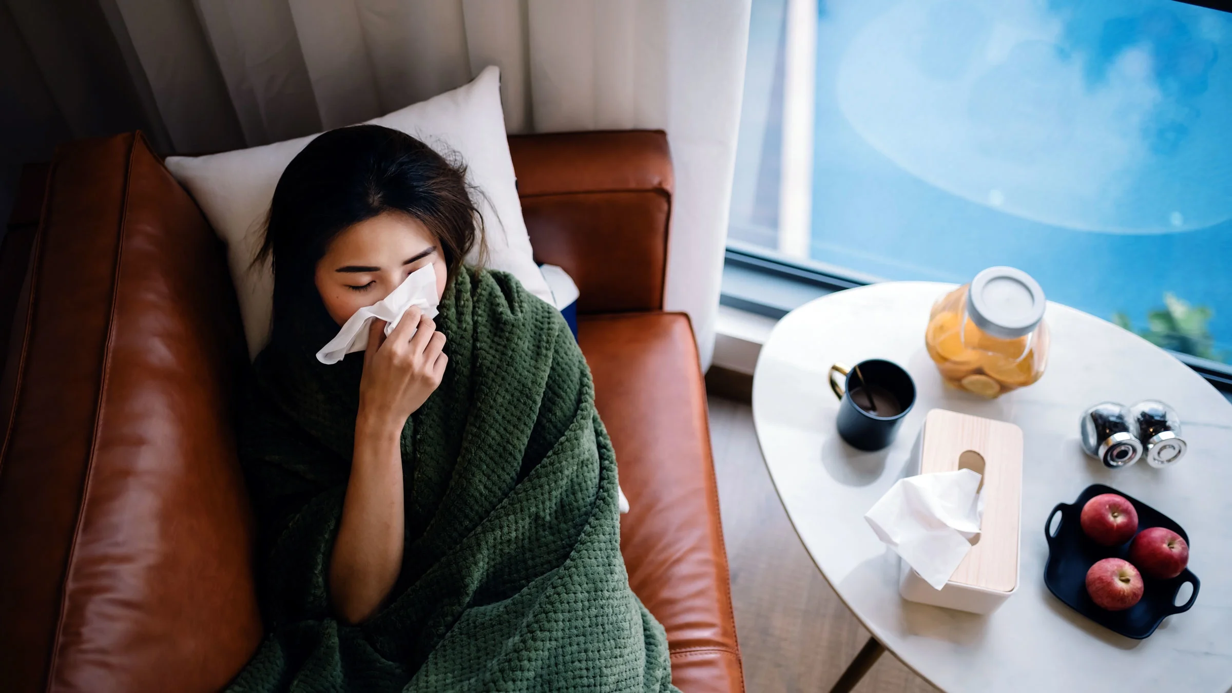 A woman is blowing her nose while lying on a sofa covered with a blanket.