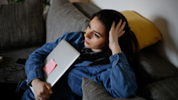 A woman looks stressed while sitting on a sofa and holding a closed laptop.
NSimages /iStock via Getty Images Plus