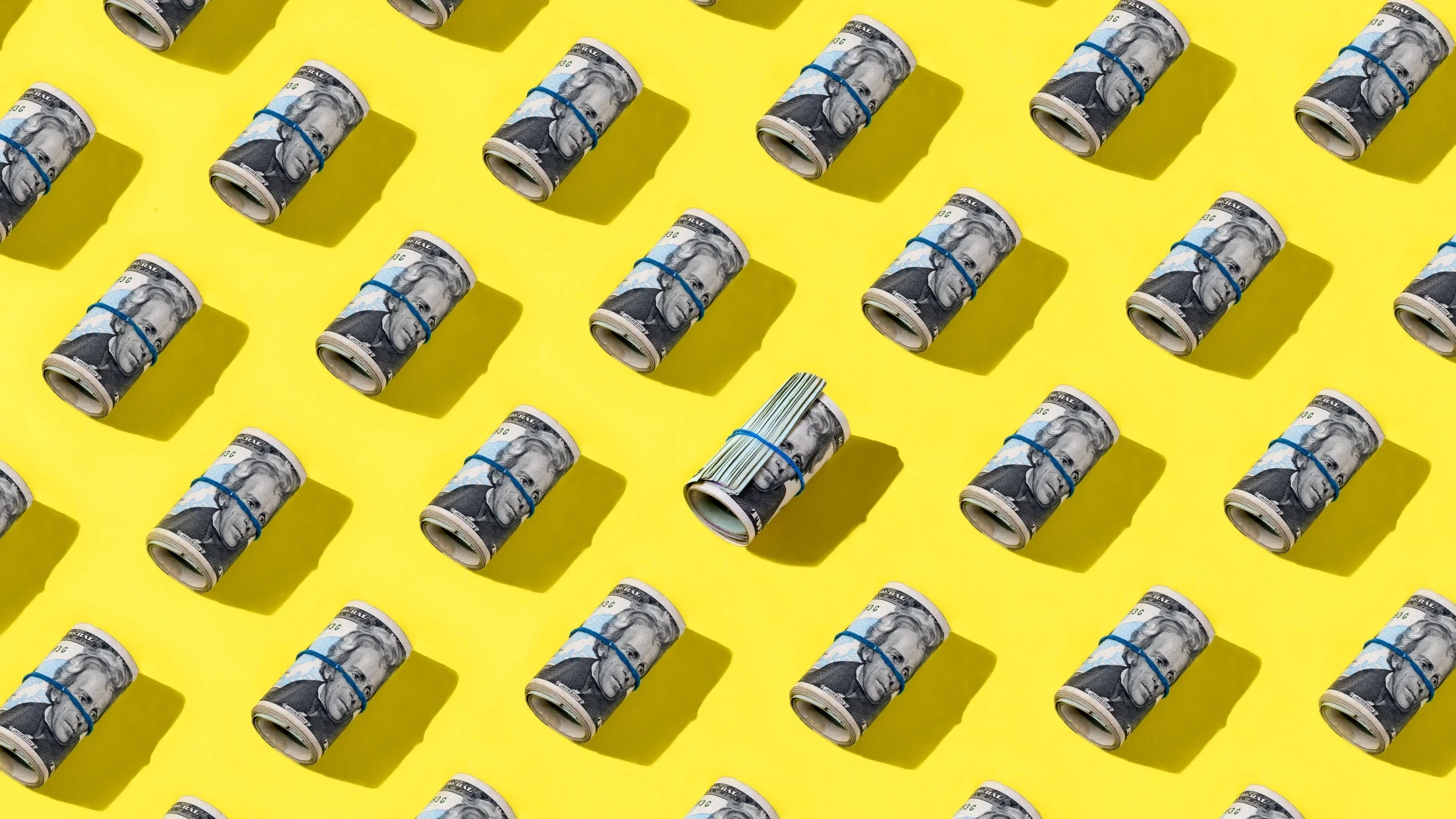 Rubber banded rolls of cash lined up diagonally in rows across the frame on a yellow background.