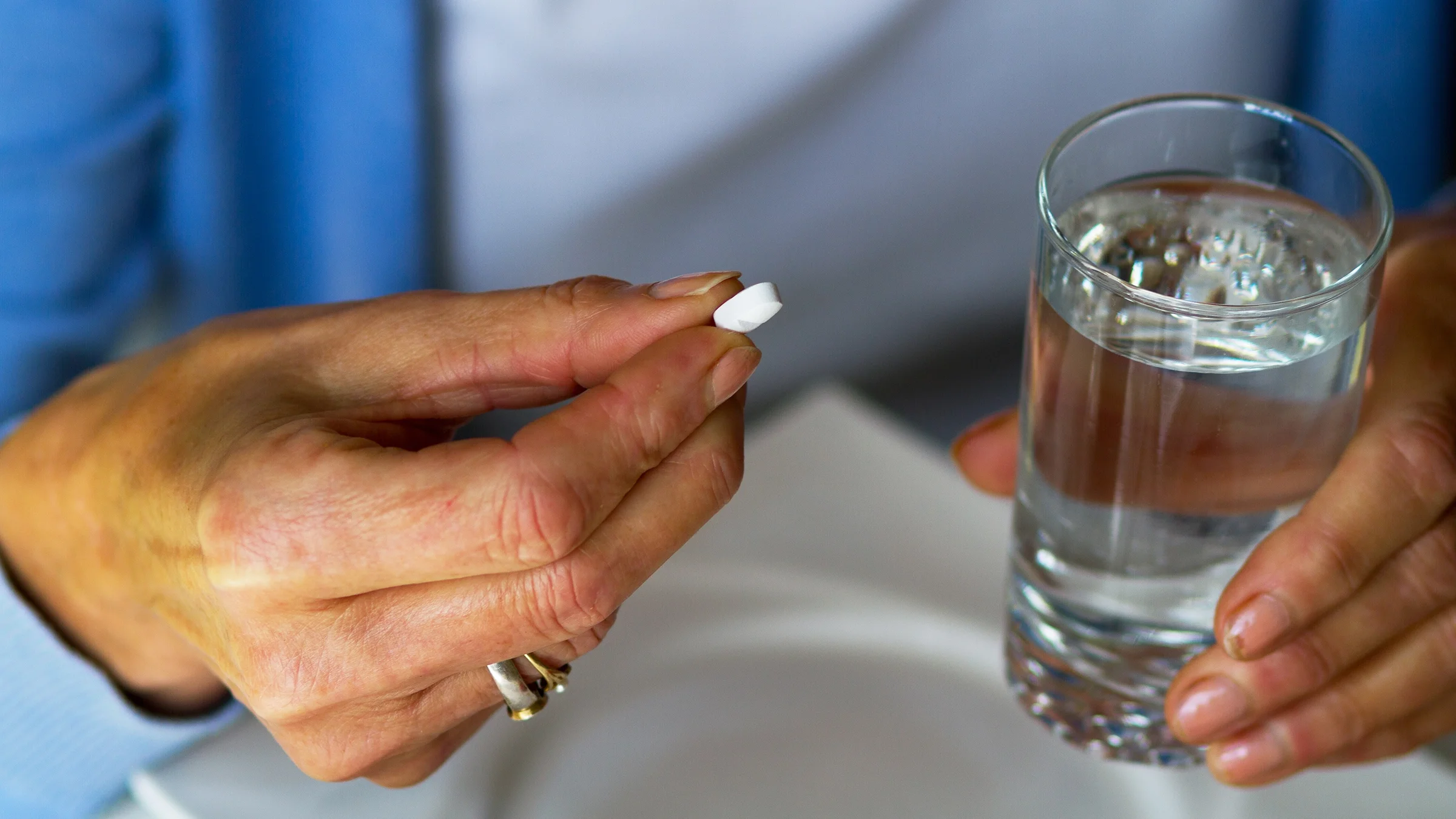 Close-up of a person holding a white pill in between their fingers. They are holding a glass of water in their other hand.