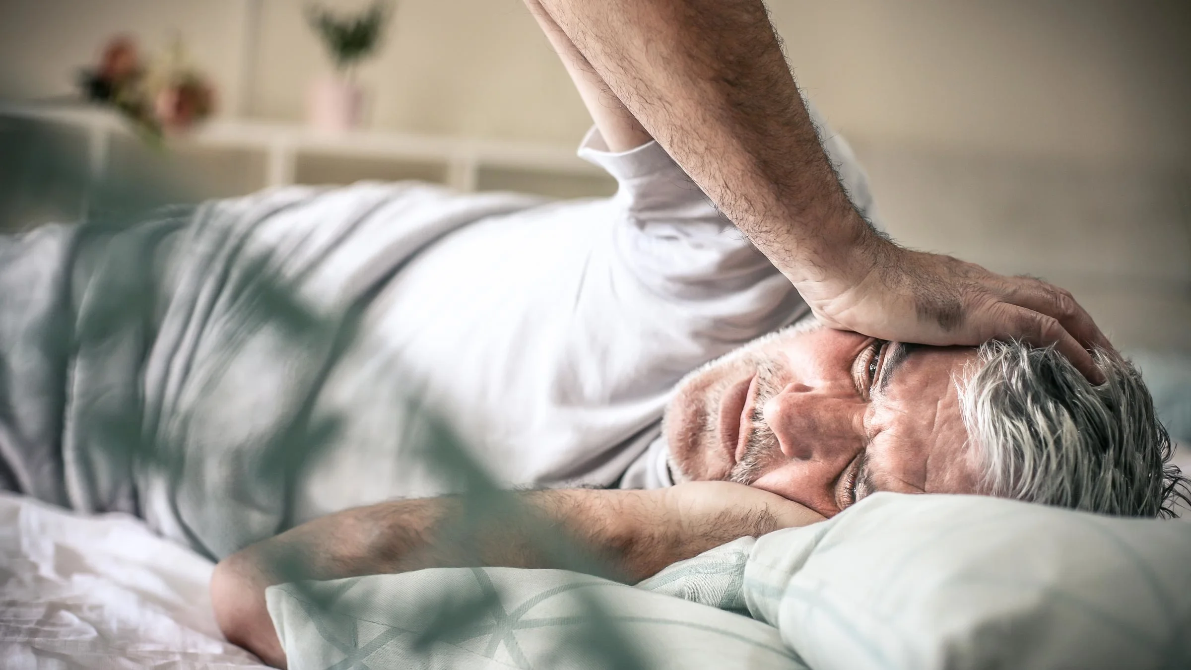 An older man lies in bed while suffering from a headache.