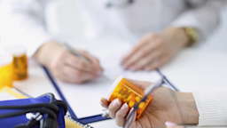 Cropped picture of a patient's hands holding a pill bottle, with a doctor taking notes in the background.
Ivan-balvan/iStock via Getty Images Plus 
