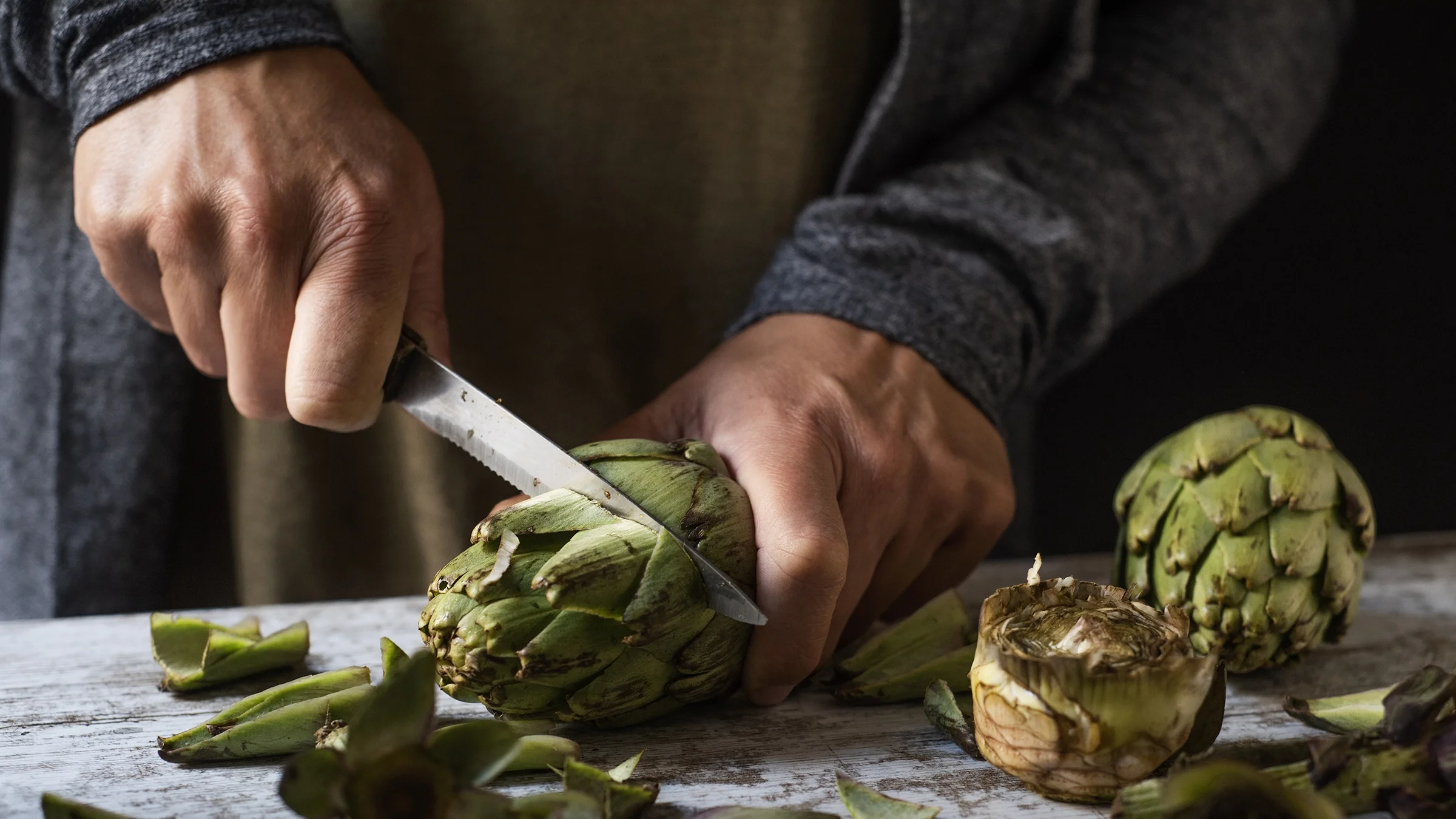 A man is cutting fresh artichokes on a rustic wooden table.