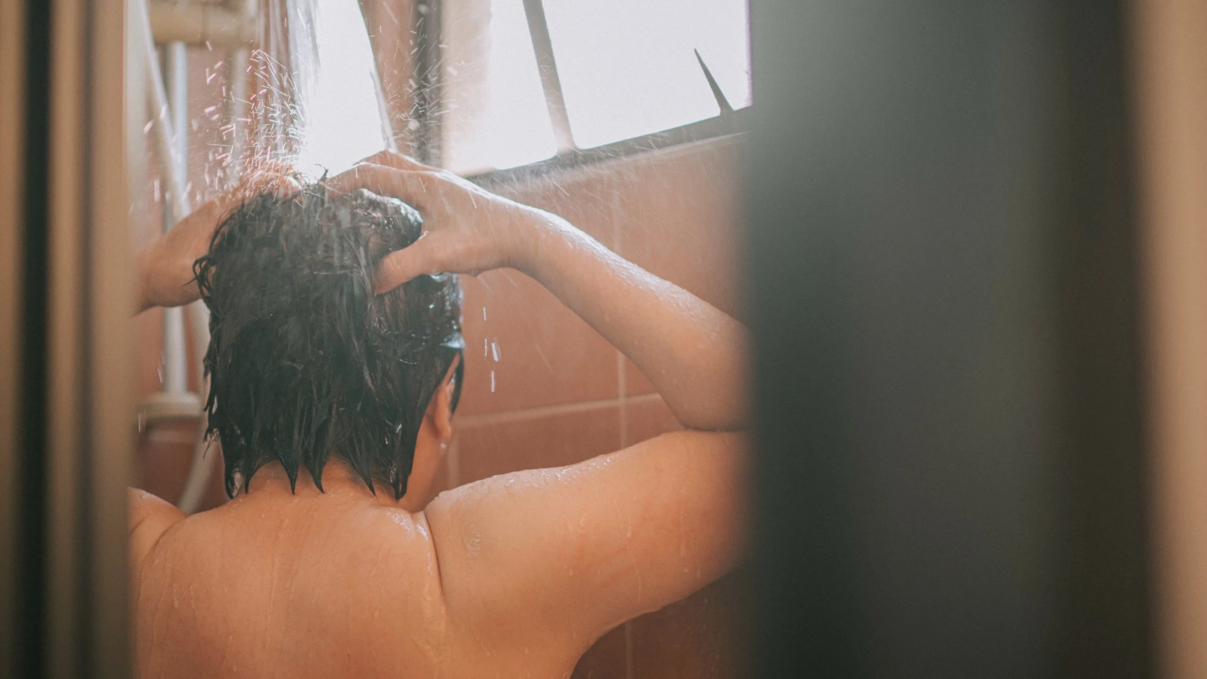 Woman in the shower washing her hair, looking at her back framed by the shower door frame on either side.
