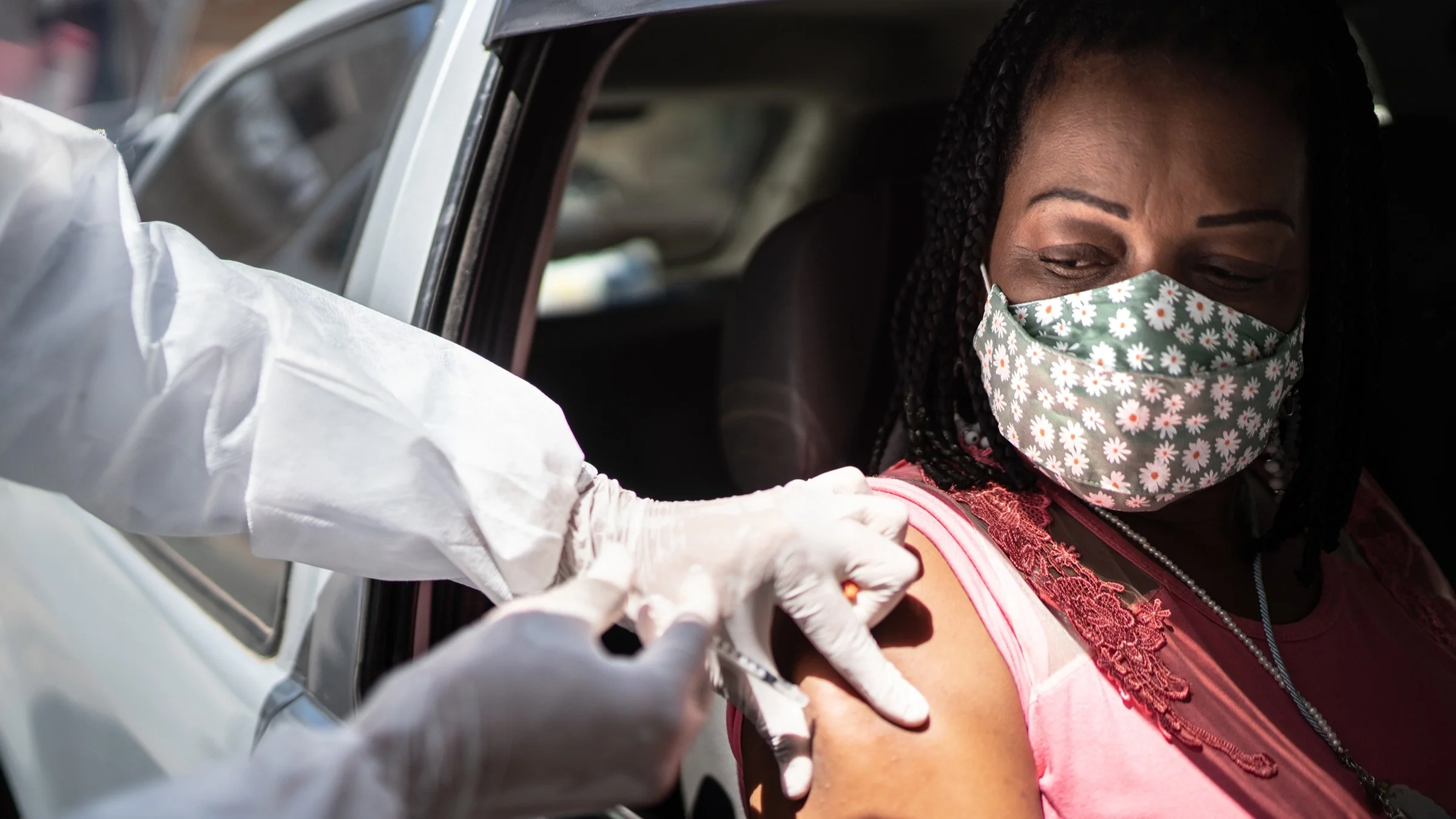 Woman in her car with a flower face mask on getting her COVID-19 vaccine from a drive-thru vaccine site.