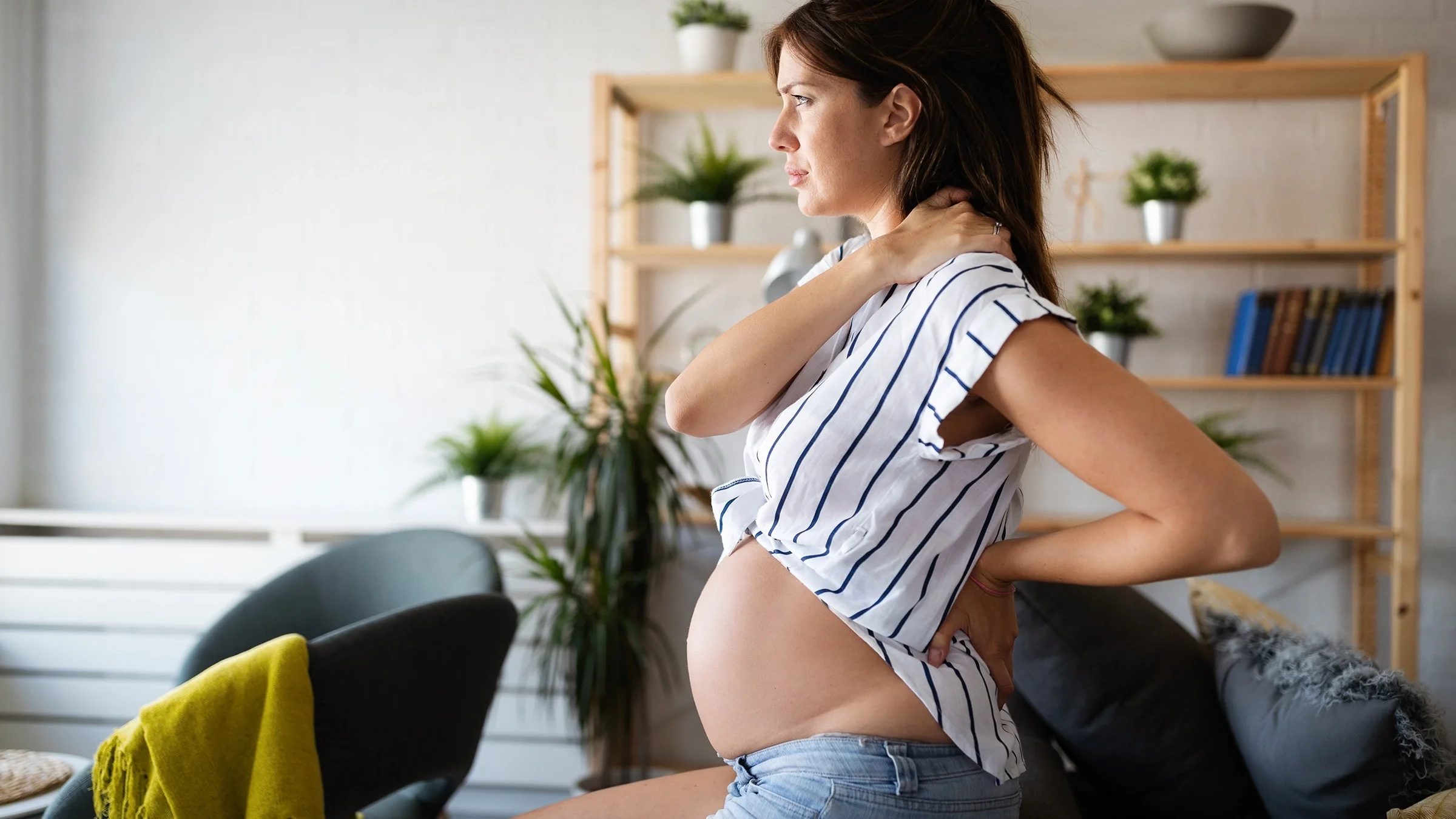 Pregnant woman sitting on the edge of the couch rubbing her sore neck.
