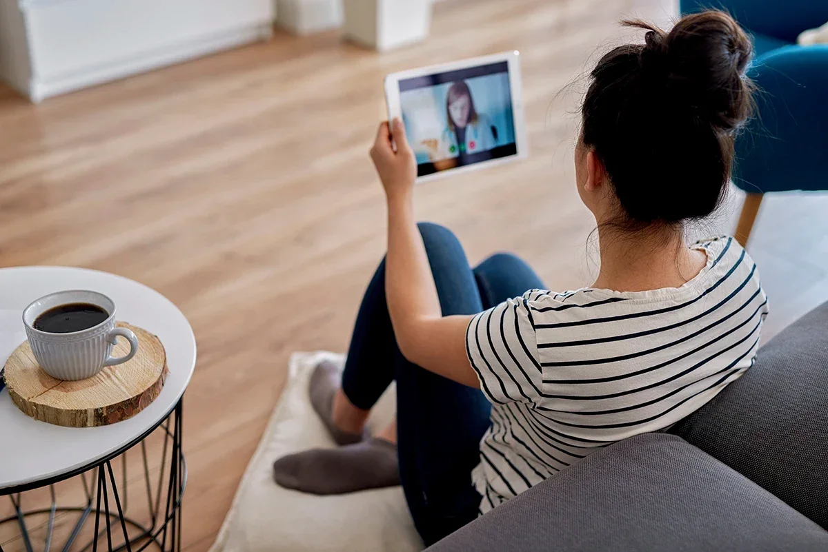 Young woman with her hair in a bun sitting in front of her couch talking to her doctor on a telehealth visit on her tablet.
gpointstudio/iStock via Getty Images