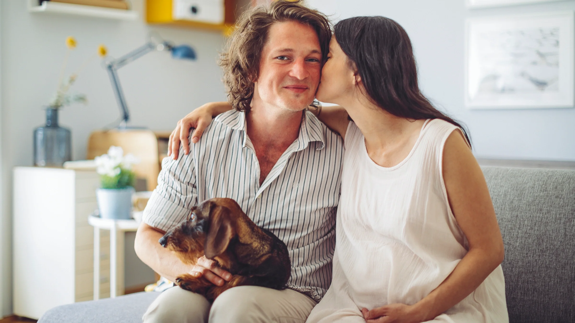 Couple posing for a photo in their house. The woman is pregnant and the man is holding their dog in his lap.