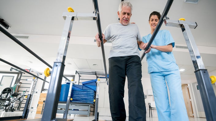 Senior man at an occupational therapist appointment. The therapist is helping him walk using the parallel bars in the equipment room.