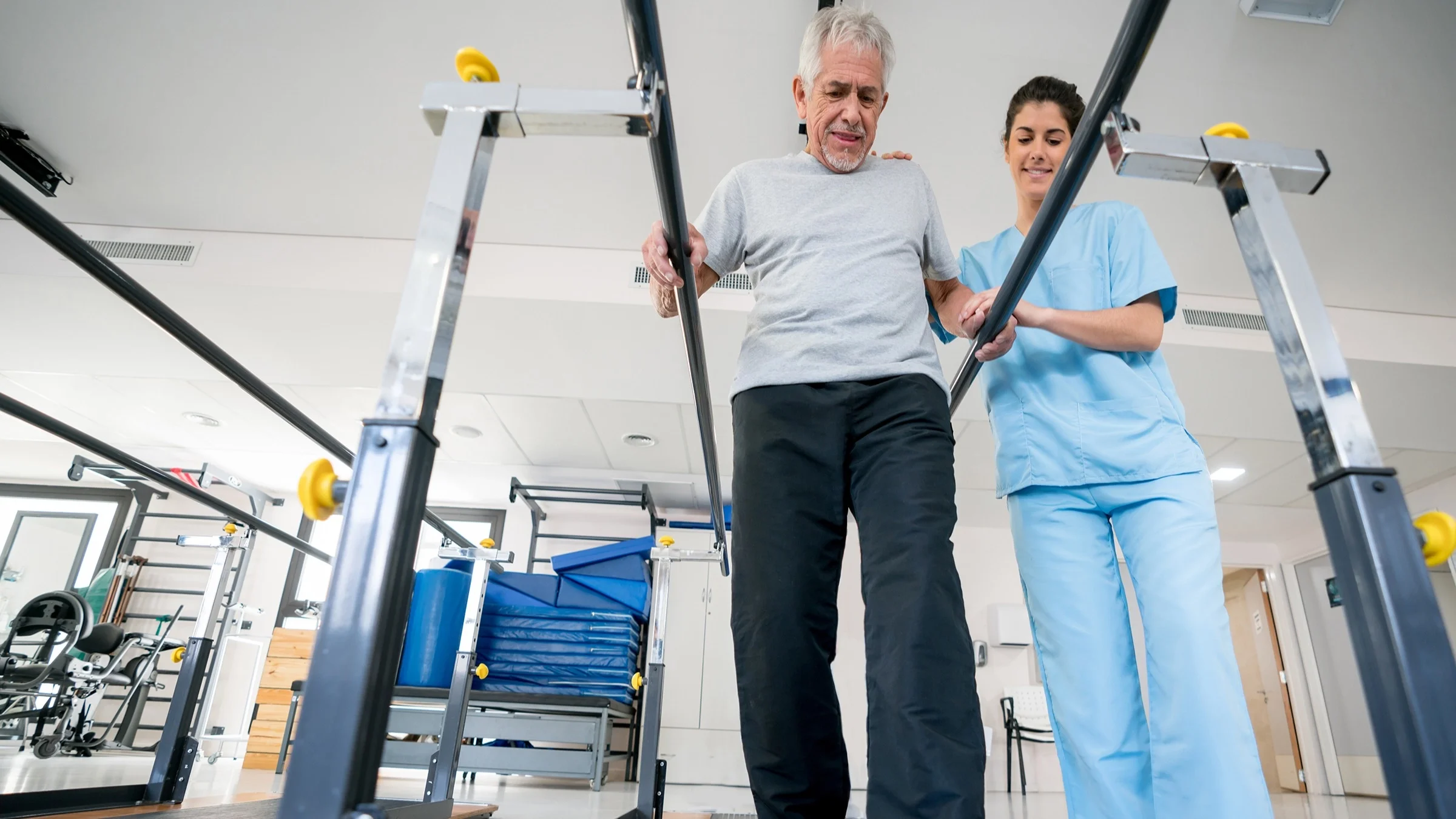 Senior man at an occupational therapist appointment. The therapist is helping him walk using the parallel bars in the equipment room.