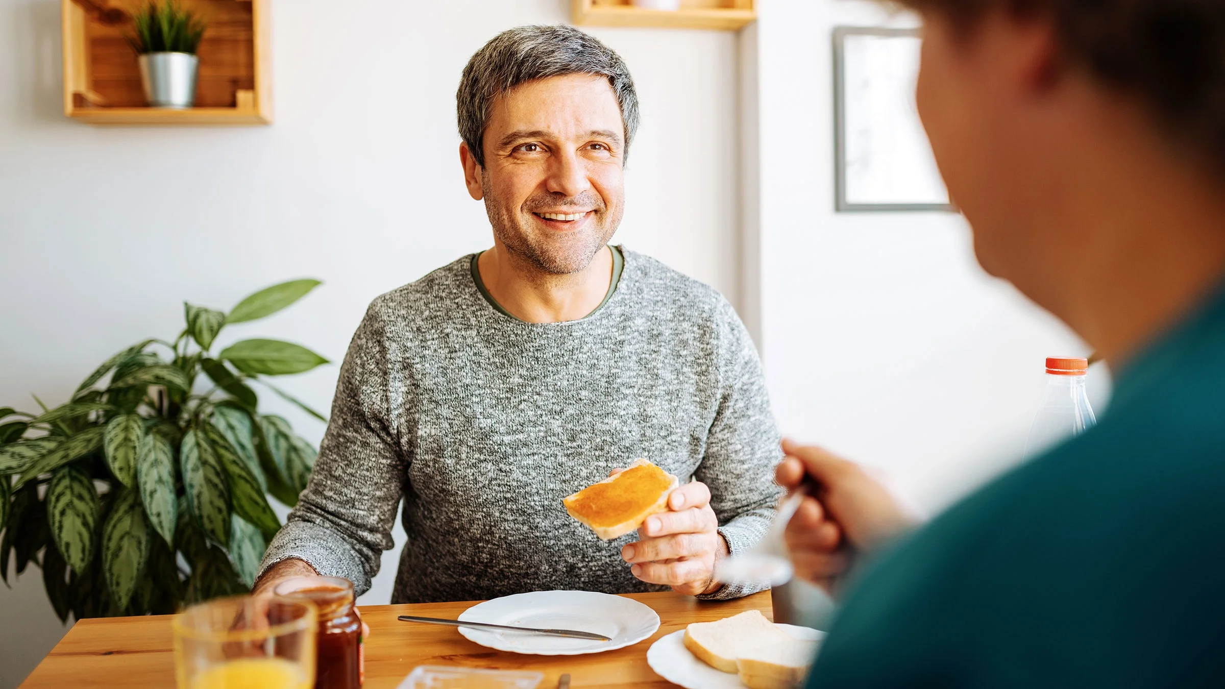 Man smiling and offering a piece of toast at a breakfast table with orange juice