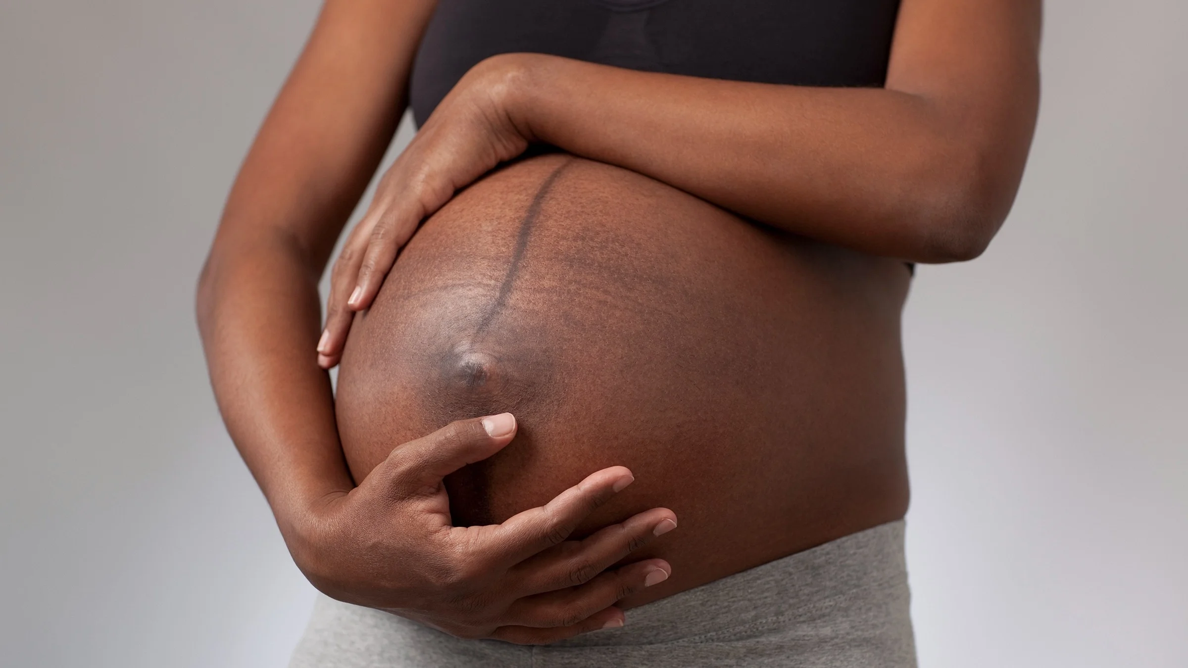 Cropped shot of a Black woman holding her pregnant belly. There is a dark vertical line down her abdomen.