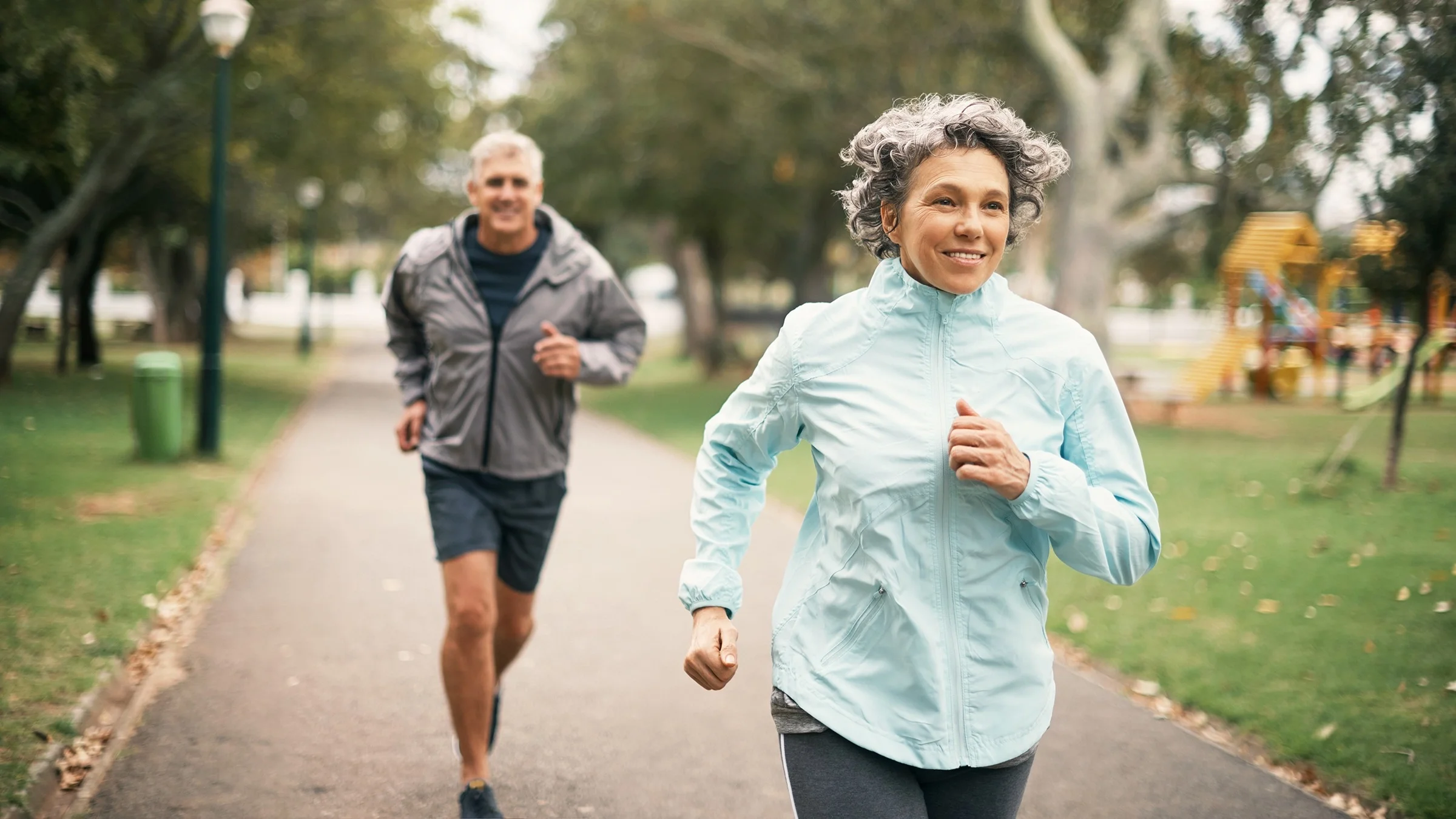 Senior couple going for a run through the park. The woman is in the front in focus in a light blue and gray workout outfit.