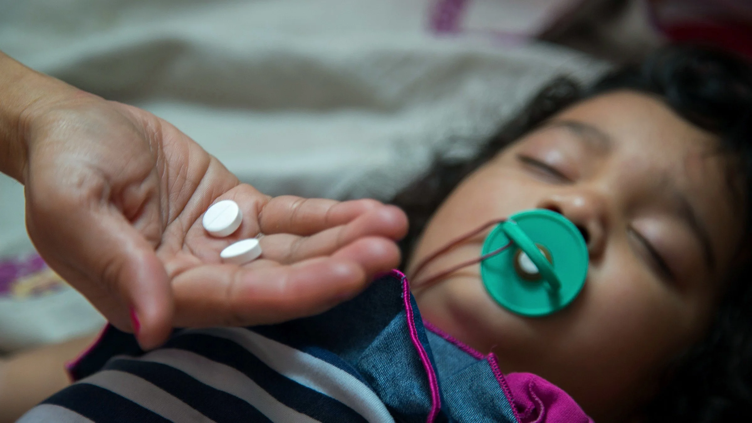 Close-up on a parent's hand holding two tablets of aspirin. You can see their sleeping baby in the background with a pacifier but they are out of focus and blurry.