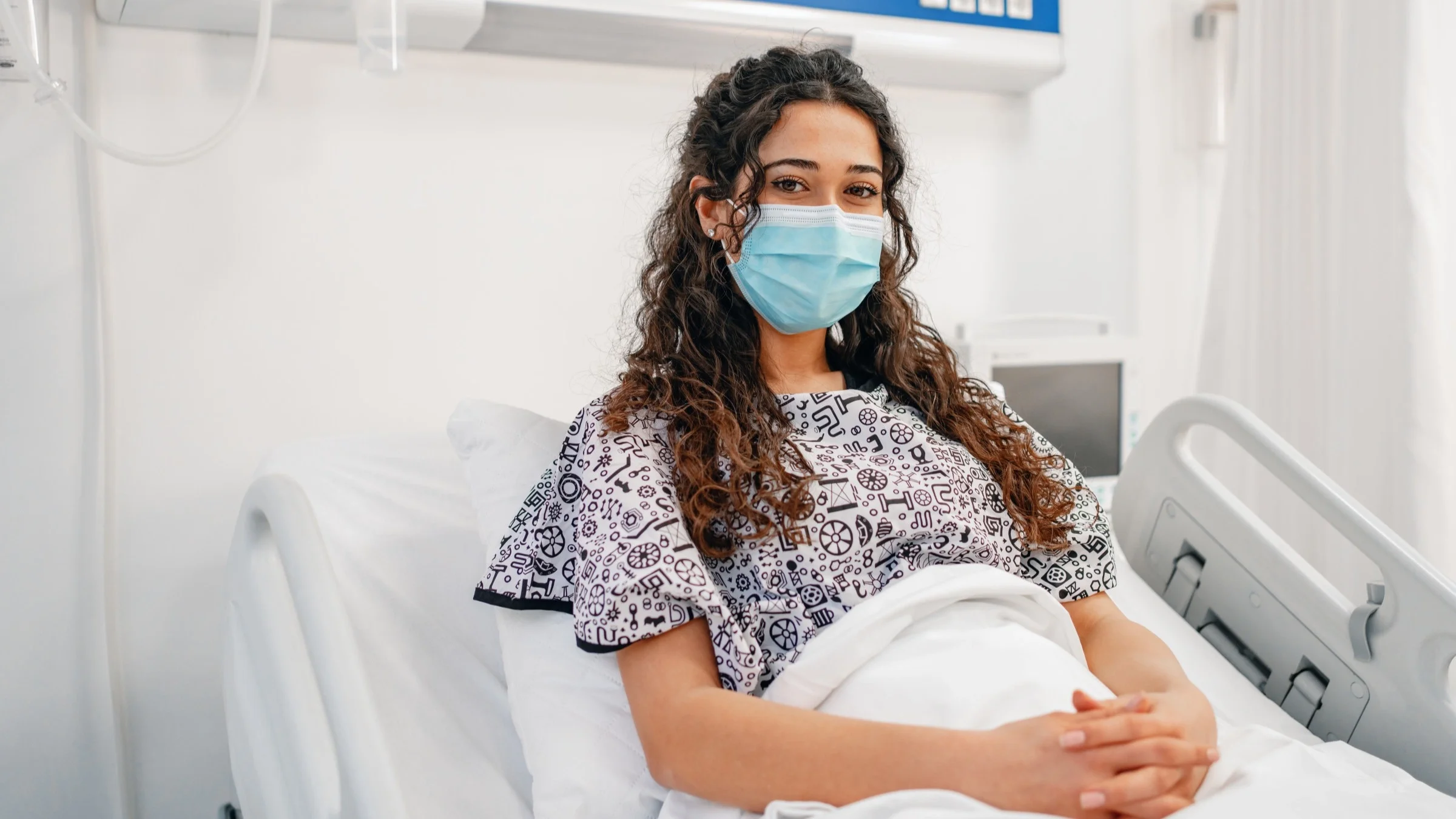 Portrait of a patient in a hospital bed. She is sitting up and wearing a medical face mask.