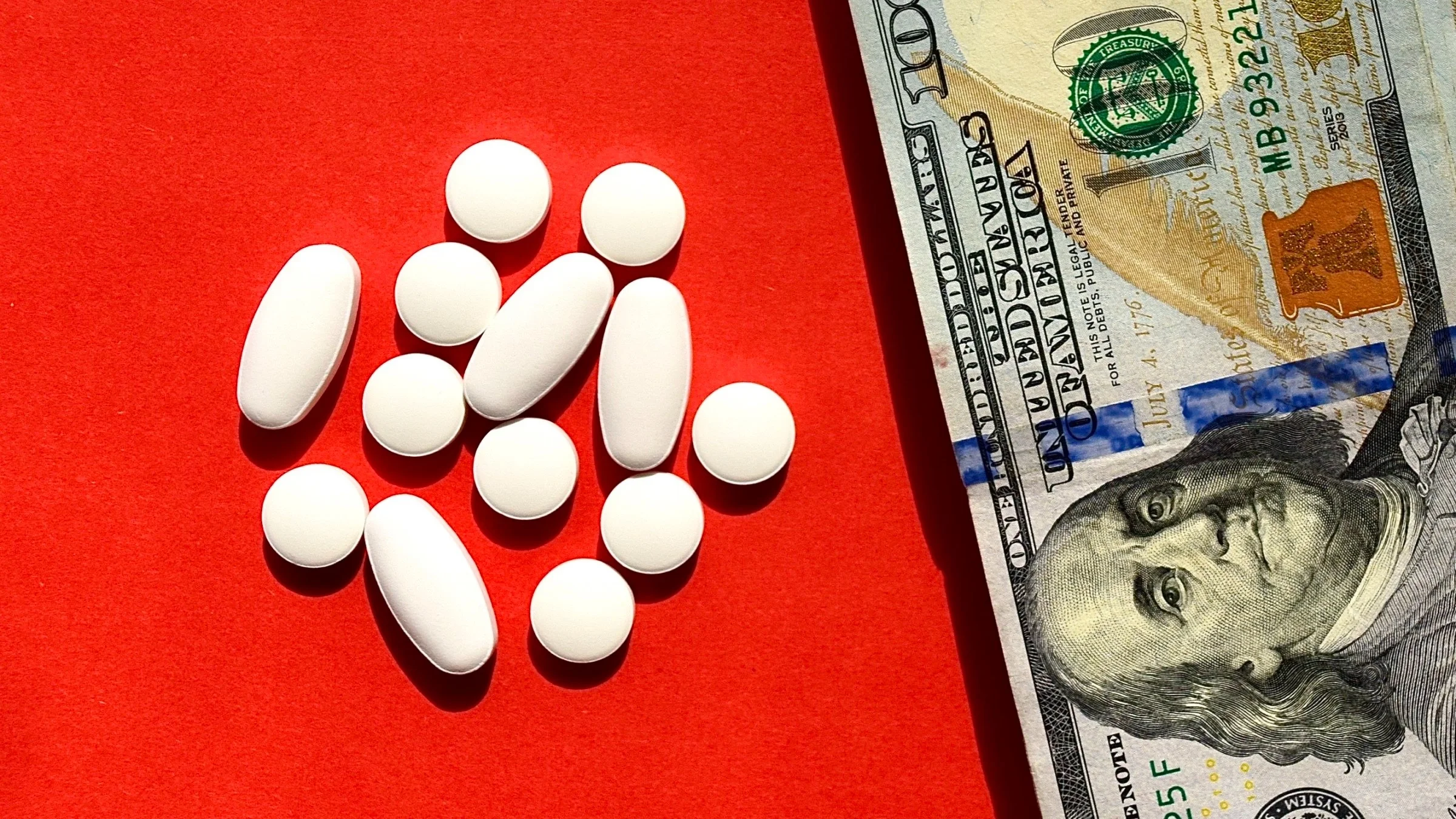 Pile of white pills of various sizes and shapes next to a hundred dollar bill on a bright red background.