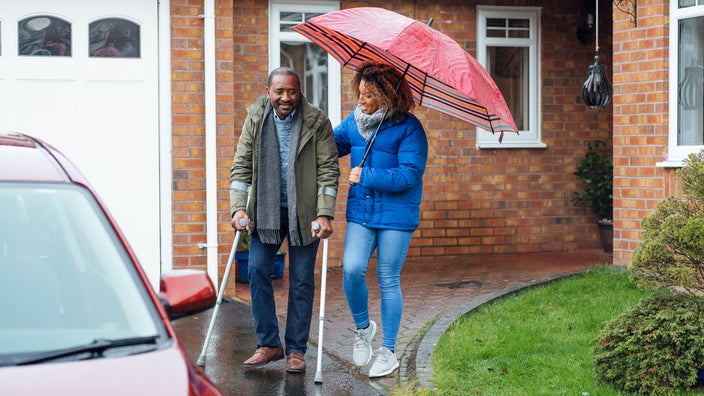 An older man is walking on crutches to a car with help from his daughter.