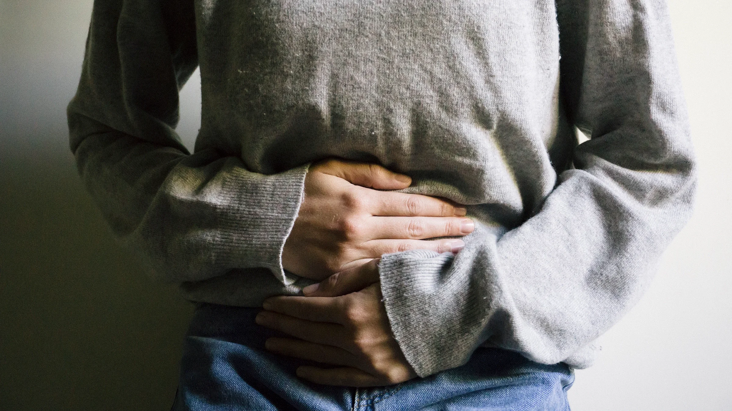 Cropped close-up of a person holding their abdomen in pain. They are wearing a light gray sweater.