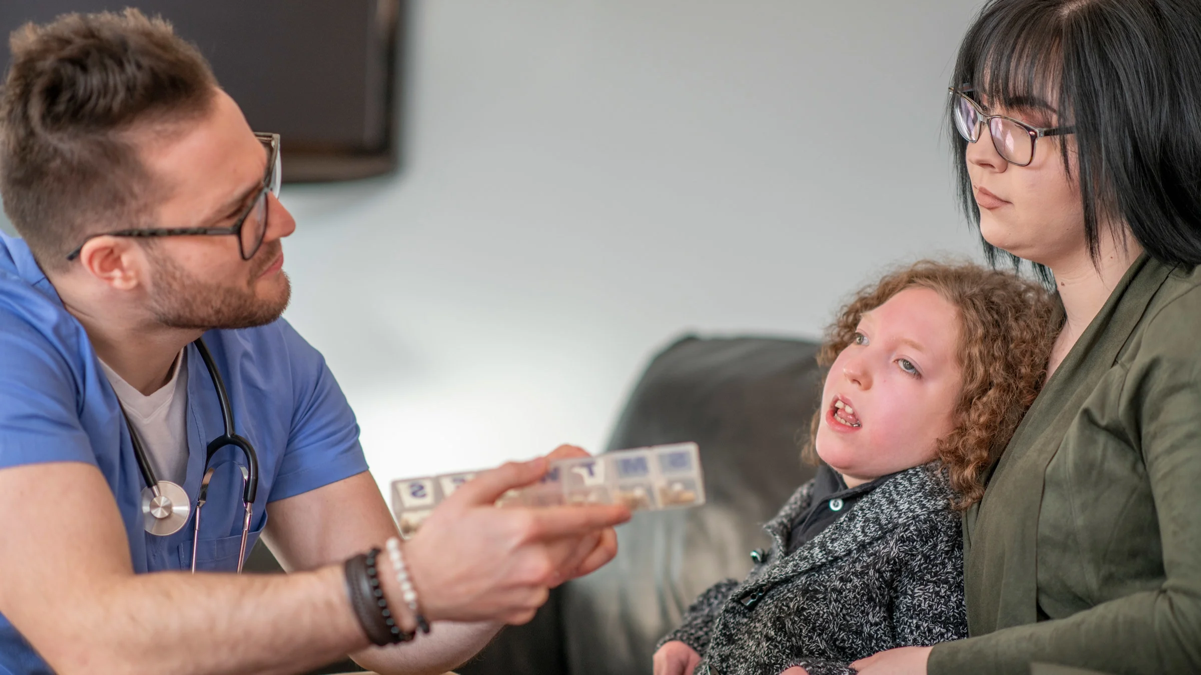 A HCP showing a pill container to a patient with cerebral palsy and their parent.