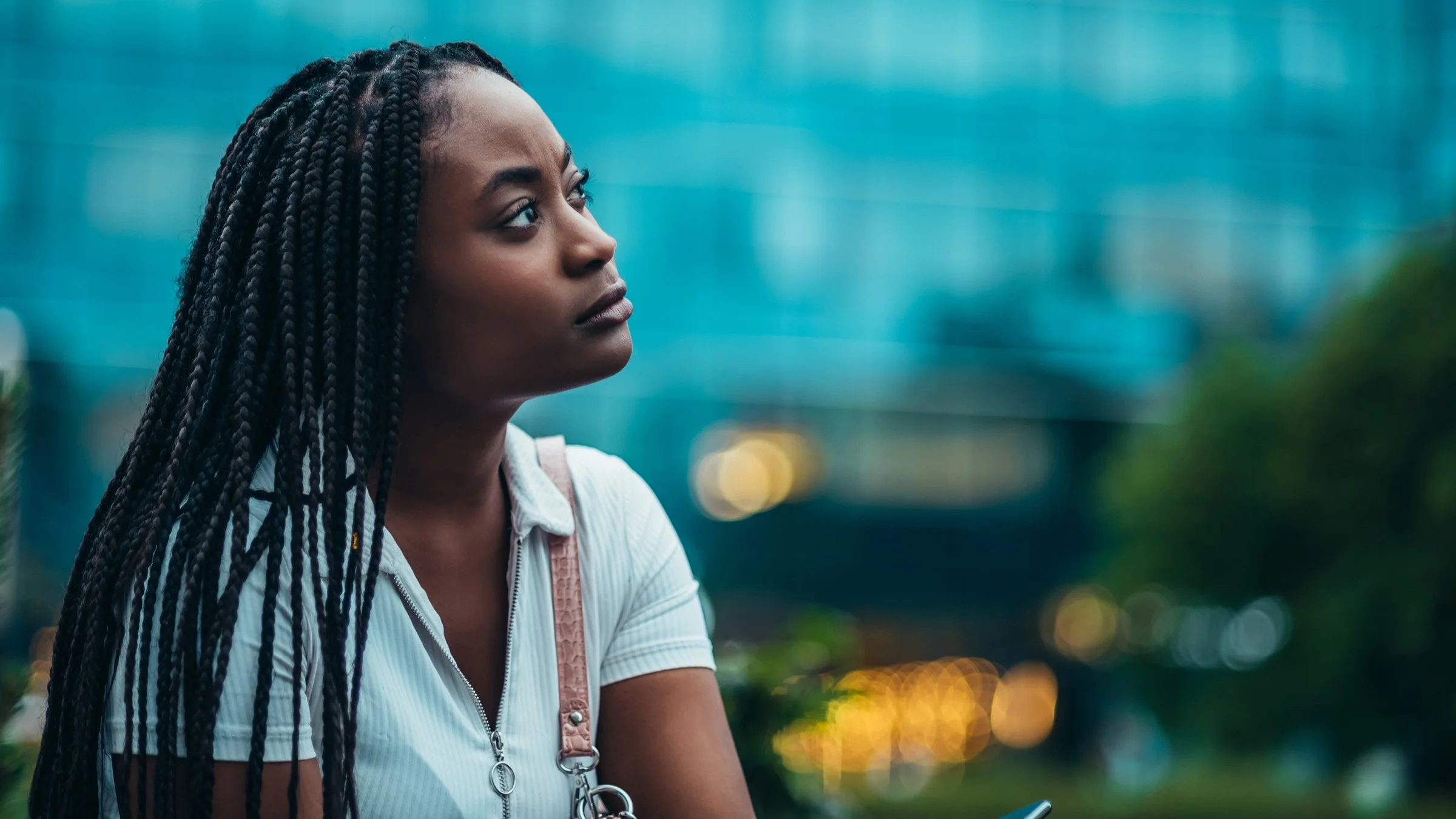 Portrait of a young beautiful woman with braids looking upward. She's surrounded by a cityscape of buildings.