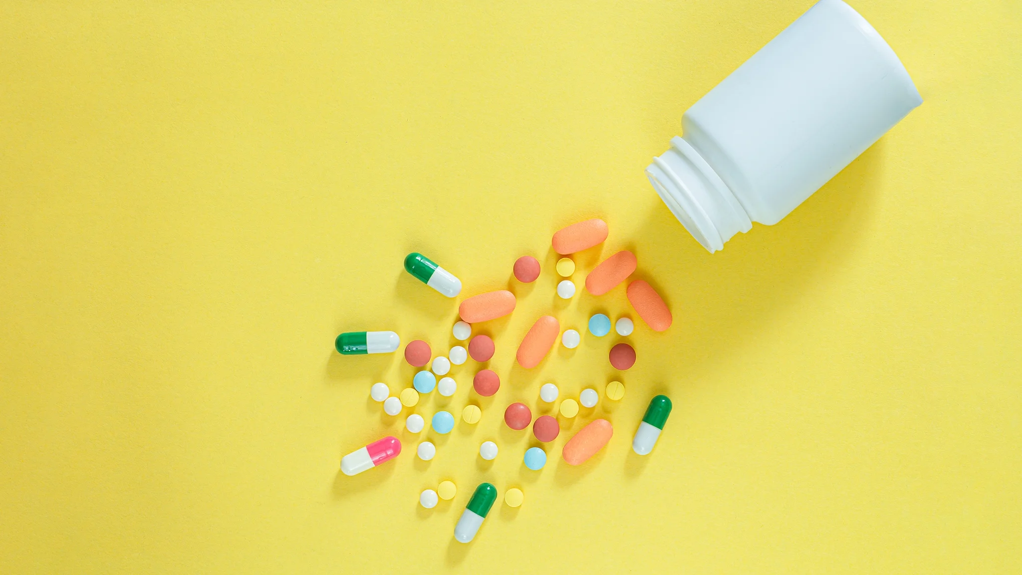 Variety of pills spill out of a generic white pill bottle on a yellow background.