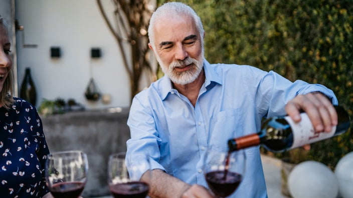 Older man with a beard pouring a glass of wine for his friend at an outdoor dinner party.