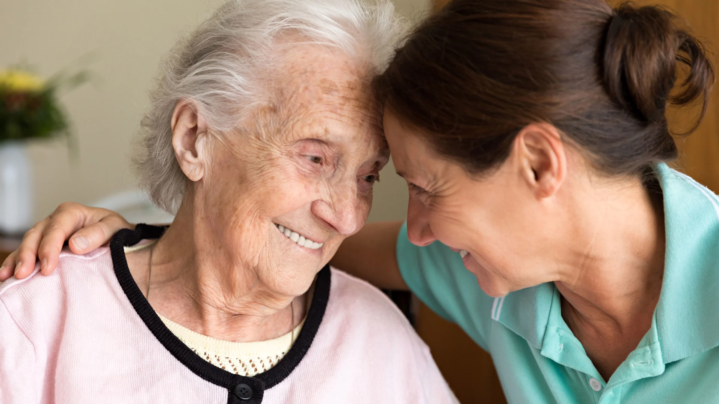A close-up portrait of an older woman and her adult daughter. They are resting their foreheads together and smiling lovingly.
