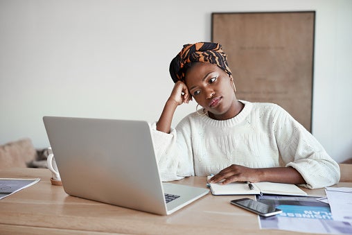 Young woman in a headwrap sitting looking sad at her laptop with notes, and papers surrounding her at her table.
