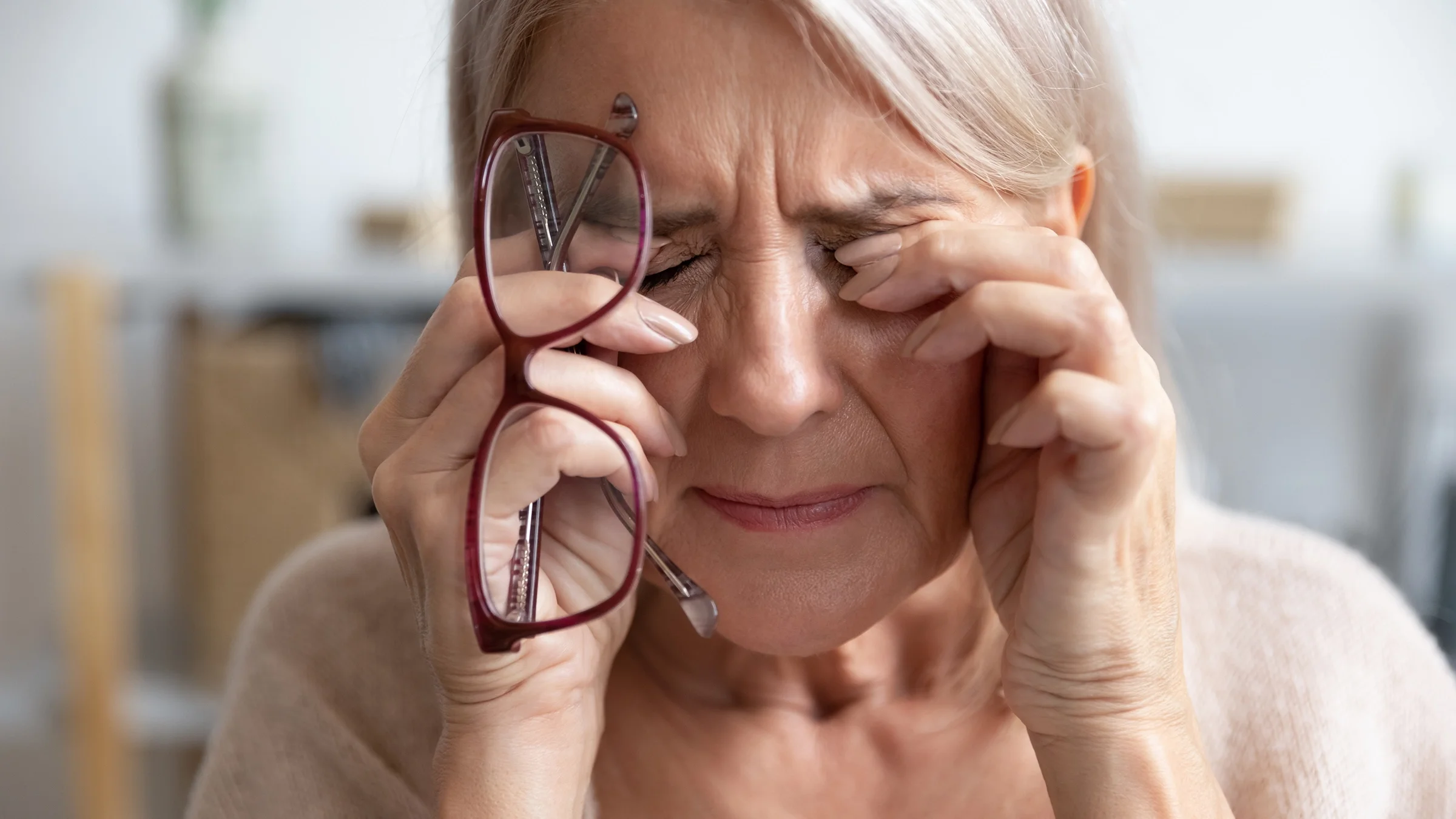 Close-up of a woman rubbing her eyes in pain while holding her glasses in her hand.