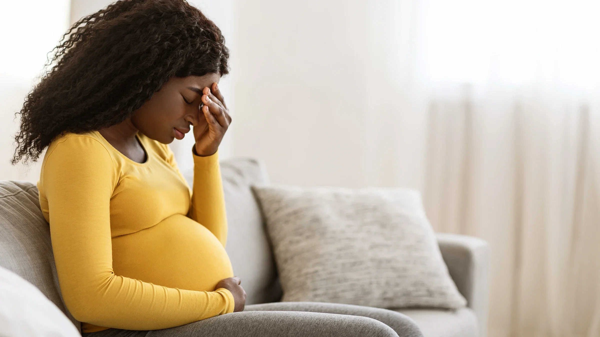 Black pregnant woman sitting on the couch with a headache. She has her hand on the middle of her forehead. She is wearing a yellow long sleeve shirt and gray yoga pants.
Prostock-Studio/iStock via Getty Images